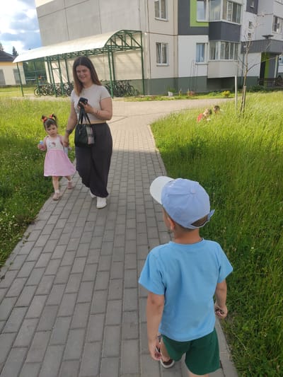 The image shows three people standing on a paved walkway next to a grassy area. In the foreground, a young girl wearing a pink dress with a "Hello Kitty" graphic is holding hands with a woman in a striped shirt and black skirt. The woman is smiling and looking towards the camera, holding a phone and a bag. Behind them, a young boy wearing a blue t-shirt and a blue baseball cap is walking away from the camera. He is wearing green shorts.
The background shows a modern apartment building with balconies and grey, white, and green facades. There is a covered area with bicycles parked underneath. The scene appears to be a residential area, possibly a courtyard. The weather seems to be sunny, with good lighting. The time of day appears to be daytime. There is some text visible on the boy's blue cap, which appears to say "SAUR". The overall mood seems relaxed and casual, suggesting a family outing or a walk in the neighborhood. The location is Navapolatsk, Belarus.