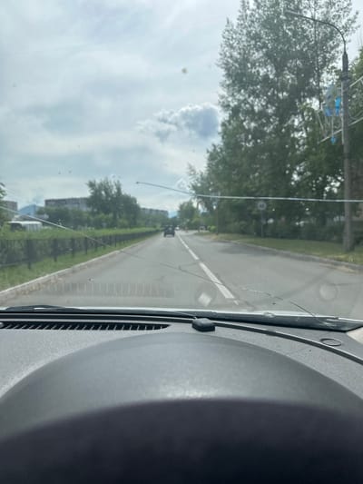 The image shows a view through a car windshield. The windshield has several cracks and is dirty. The car is on a paved road with white lane markings. On the left side of the road, there is a metal fence and then some buildings in the distance. On the right side of the road, there are trees and a lamppost with a speed limit sign that appears to indicate 60. The sky is overcast with clouds. The dashboard of the car is visible in the foreground. The location appears to be Sayanogorsk, Russia, based on the context provided.