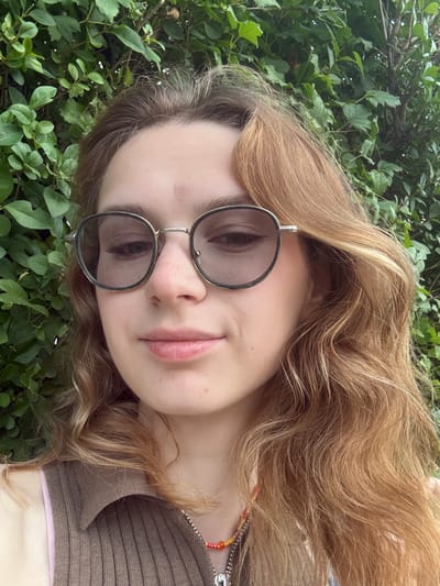 The image displays a close-up, chest-up portrait of a young individual with fair skin and long, wavy, light brown hair, exhibiting some lighter, possibly auburn, highlights. The hair frames the face and falls over the shoulders. The individual is wearing round-framed sunglasses with dark, subtly tinted lenses and metallic, silver-toned frames, obscuring the eyes. A small reddish blemish is visible on the upper forehead, and another is present on the lower right cheek. The nose is straight, and the lips are naturally pink, appearing full and slightly parted with a neutral to subtly smiling expression.
The individual is dressed in a brown, ribbed, collared top, likely a knit material, with a visible zipper detail on the right side. A pale pink strap or edge of another garment is visible underneath the brown top on the left shoulder area. Around the neck, a layered necklace is worn, featuring one strand of small, colorful beads (orange, yellow, pink) and another metallic chain with an indistinct silver-colored pendant.
The background is entirely filled with dense, vibrant green foliage, comprising numerous leaves and some visible branches, indicating a thick hedge or bush. The setting is outdoors with natural light. No specific urban or geographical landmarks are discernible within the frame to confirm a location such as Kragujevac, Serbia.
