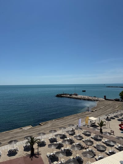 The image depicts a beach resort area in Kavarna, Bulgaria, seen from an elevated perspective, potentially from a high building. The scene is primarily focused on a stretch of coastline with a blue sea on the left, gradually transitioning to a clear, light blue sky. A pier extends into the sea, bordered by rock structures, likely serving as a breakwater. To the right of the sea, a sandy beach is arranged with rows of thatched umbrellas and sunbeds. A wooden boardwalk runs along the edge of the beach, parallel to the shoreline. Several palm trees are strategically planted amongst the umbrellas, providing shade. Flags and banners are visible near the edge of the beach, suggesting promotional or branding elements. The overall impression is a tranquil beach scene during a clear day.