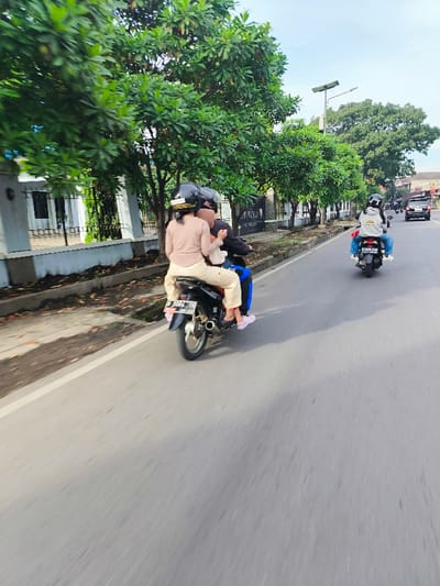 The image shows a woman riding a motorcycle on a paved road in the city of Kecamatan Margahayu, Indonesia (latitude: -6.977, longitude: 107.561). She is wearing a helmet and a beige shirt.  A second person is sitting behind her, and a third person is sitting behind them, holding onto the woman. The woman is riding the motorcycle in a residential neighborhood with a single lane road lined with trees. There are houses on one side of the road and a white curb. The motorcycle is moving to the right of the frame. Another motorcycle, with a person wearing a grey jacket, is in the distance moving in the same direction.  The sky is clear, and the weather appears sunny.  There are no cars in the background.
