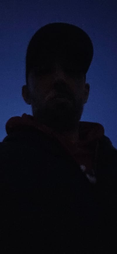 The image displays a person captured from a low-angle perspective, predominantly in silhouette against a dark blue background. The individual's head and upper torso are visible, with the head positioned towards the upper right of the frame, tilted downwards or viewed from below. Although most of the face is obscured by deep shadow, faint outlines of a nose, mouth, and chin can be discerned. The person appears to be wearing dark clothing, which blends into the shadow.
The background is a uniform, deep, dark blue, suggesting either a featureless surface like a wall or fabric, or possibly a night or deep twilight sky. The lighting conditions are extremely low, creating a dramatic contrast between the dark figure and the dark blue setting. No specific activity, emotions, or identifiable objects are visible, nor is any text present. The scene conveys a sense of stillness or observation in very dim light, with the location context being South Norfolk, United Kingdom.