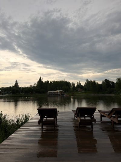 The image depicts a serene, overcast day by a body of water. In the foreground, a wooden deck is wet, reflecting the muted light. Several empty wooden lounge chairs are positioned on the deck, facing the water. Reeds and green vegetation grow along the edge of the deck.

The water of the lake or river is calm, with soft reflections of the trees and sky. Across the water, a line of dense green trees forms the background. A long, low building with a light-colored roof is visible nestled among the trees on the far bank. Further to the left, a playground structure can be seen.

The sky is filled with heavy, grey clouds, suggesting a recent or impending rain. The overall atmosphere is one of tranquility and quietude. There are no people visible in the image. The scene appears to be a public park or recreational area, possibly a resort, situated in or near the city of Chusovoy, Russia, given the context provided.