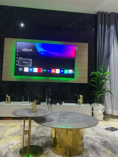 A contemporary living room features a wall-mounted television with a vibrant display and ambient green backlighting. A white console table underneath the TV holds gaming equipment and decorative gold figurines. Adjacent to the console, a striking gold and marble tiered coffee table sits on a textured rug. A potted plant and decorative white vase are placed atop the console table. Gray drapes adorn the wall behind, contributing to a modern aesthetic.