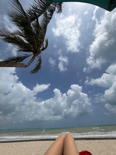This is a low-angle, eye-level shot of a beach scene in Bang Muang, Thailand. In the foreground, a person's legs are visible, suggesting they are lying down on a beach towel. The beach itself is sandy with scattered small rocks and shells. A concrete retaining wall separates the sand from the ocean. The ocean water is choppy with moderate waves and appears to be a murky green-blue color. The sky is a bright blue with scattered white and grey clouds, with a portion of a dark grey cloud bank visible in the distance, hinting at potential rain. A palm tree leans into the frame from the upper left corner, its fronds swaying in the wind. A small portion of a turquoise beach umbrella is visible at the top of the frame.