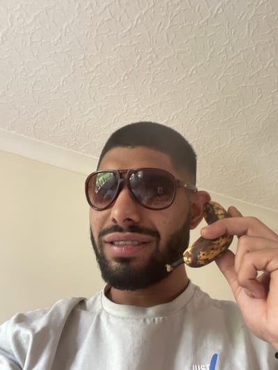 A close-up, angled shot depicts a person with dark hair, a beard, and sunglasses, holding a ripe, slightly browned banana near their ear as if it were a telephone. The person is wearing a light-colored t-shirt. The background is a textured white surface, possibly a wall. The location is not discernible from the image.