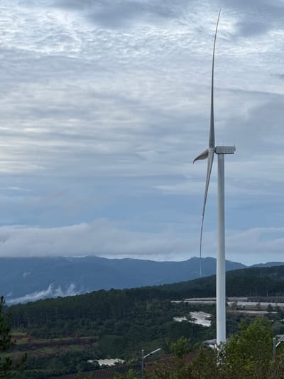 A landscape in Phát Chi, Vietnam, features a wind turbine with two visible blades, against a backdrop of cloudy skies and mountainous terrain. The turbine stands to the left side of the frame. Patches of forested area and what appear to be agricultural coverings can be seen along the slopes of the distant mountains.