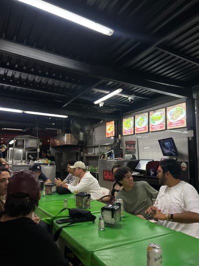 The image captures a casual, brightly lit indoor or semi-outdoor eatery, likely a taqueria, in Mazatlán, Mexico. Several young men are gathered around a bright green table, engaged in conversation and appearing to dine or wait for their orders.
In the foreground, a person wearing a maroon baseball cap is partially visible. Further back, a man in a white t-shirt and glasses is seated at the table, leaning forward. Across from him, another man in a light green t-shirt looks out towards the left. On the other side of the table, a man in a white and black striped shirt and a white baseball cap is visible, alongside another individual wearing a dark baseball cap with a visible "NY" logo.
The green table is covered with a plastic cloth and holds a silver can with "JACK D" (Jack Daniel's) visible, other beverage containers, a napkin dispenser, and salt shakers.
Above the dining area, several digital menu boards display various food items with images and text, including "TACO DE ASADA DE MAIZ," "VAMPIRO," "QUESADILLAS," and "ORDEN DE CARNE ASADA," reinforcing the Mexican food theme. A small television screen is mounted on the wall. The ceiling features an industrial corrugated metal structure with bright fluorescent strip lights and a large industrial fan providing circulation. Other details include security cameras and a visible sign that reads "CAJA" (cash register) and "CUALPA."
The artificial lighting suggests the scene takes place in the evening or at night. The atmosphere appears relaxed and social, typical of a casual dining experience.