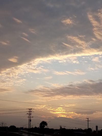 The image depicts a sky at sunset over what appears to be a highway. The sky has layers of clouds, ranging from darker, gray-toned clouds at the top to orange and yellow-tinged clouds near the horizon where the sun is setting. Below the sky, there is a road with vegetation and infrastructure, including electrical poles and lines, and silhouettes of trees. A few vehicles can be seen on the road. The information provided states that this scene is in El Dátil, Venezuela, but this can't be confirmed from the visual data.