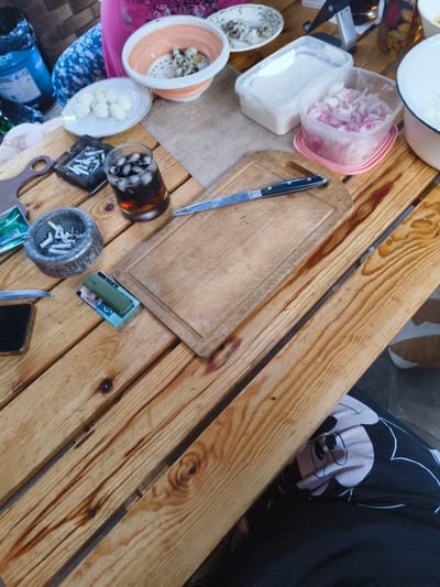 The image depicts an outdoor gathering on a wooden table, possibly in Востец Снт, Russia. A glass of dark liquid with ice and a cigarette ashtray filled with butts are on the table, alongside a cutting board with a knife. There are also containers of chopped red onion and what appears to be a white substance, possibly sour cream or yogurt, along with a plate of boiled quail eggs. A person's lap with a Mickey Mouse pattern on their clothing is visible in the foreground. The setting suggests a casual meal or picnic.