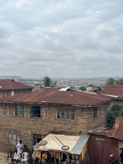 This image depicts a view of the city of Abeokuta, Nigeria, from an elevated perspective, likely a rooftop or high window. The foreground is dominated by the weathered, rusty corrugated iron roofs of several buildings, characteristic of the urban landscape. The sky above is overcast with a thick layer of grey clouds, suggesting a humid or rainy day.

In the middle ground, the architecture is a mix of brick and concrete structures with more corrugated iron roofing. Some buildings have multiple stories with visible windows. In the immediate foreground, on the right side of the image, is a small market stall with various items displayed for sale. A woman with a white headscarf is visible near the stall, and a person with headphones is also present. A person in a white t-shirt and dark pants stands near some chairs in the left foreground, interacting with others who are seated.

The background reveals a sprawling cityscape with a dense concentration of buildings, receding into the hazy distance. The overall impression is one of a densely populated urban environment with a utilitarian and somewhat aged aesthetic. The scene is typical of many towns and cities in West Africa, where such roofing materials and building styles are common.