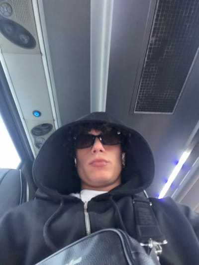 The scene depicts a young adult male inside the passenger compartment of a bus, located in the city of Nesher, Israel. The male subject, positioned centrally, is looking directly at the camera. He has dark, curly hair visible beneath a black hooded sweatshirt with a zipper, worn over a white t-shirt. He is wearing dark rectangular sunglasses and white wireless earbuds in both ears, with a silver-colored chain visible around his neck. A black shoulder bag with a wide strap is draped across his chest. The bus interior features a light grey ceiling with overhead panels containing circular vents or lights, a single blue LED indicator light, and a rectangular air conditioning vent with a grid pattern. A vertical support pole or handrail is visible towards the right side of the frame. A dark-colored seat with light piping is visible behind the subject's left shoulder, and bright ambient light enters through a window on the left.