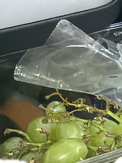 A bunch of green grapes is in a clear plastic container. The container is on a dark surface. The grapes have brown stems and some have small brown spots on their skin. The background is blurry, but it appears to be indoors. There is a grey surface above the container, and a brown surface below it. The image does not provide any information about the location of Löftaskog, Sweden.