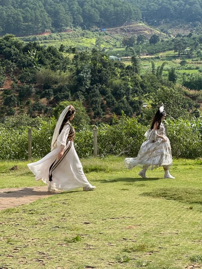 Two individuals are walking on a grassy area in an outdoor environment. The person on the left is wearing a white, floor-length dress with a veil and ornate headwear. They have a sash with black and gold detailing around their waist. The person on the right is wearing a light-colored, ornate dress with a ruffled design and a feather headpiece. Both individuals are wearing white sneakers. They are walking from left to right in the frame. The background consists of lush green vegetation, including trees and bushes, with a sloping hillside covered in trees and some scattered structures in the distance. A chain-link fence runs horizontally behind the individuals. The ground is covered in green grass with patches of dry earth and scattered small plants. The lighting suggests it is daytime. The location is identified as Thôn 5, Vietnam.