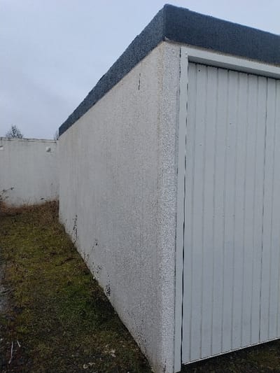 The scene in Bitburg, Germany, depicts a standalone, single-story utility building or garage under an overcast sky. The structure features exterior walls finished with a light-colored, granular texture, possibly pebbledash or coarse stucco, showing some minor staining or discoloration. A dark, flat roof with a slight overhang caps the building. On the right side of the image, a white, vertically corrugated metal roll-up door is visible, framed by white trim. The ground immediately surrounding the building consists of sparse, patchy green and brown grass mixed with bare earth. In the background, to the left, a lower, parallel white wall is discernible, with the upper branches of a leafless tree extending into the gray sky above it. No people, active subjects, or specific interactions are present in the image.