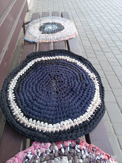 Three handmade, circular braided rag rugs are displayed on a wooden bench. The rug in the foreground is predominantly dark blue with concentric rings of black, white, and light beige. The middle rug is also circular and appears to be made from various colored fabric scraps, primarily white, blue, and black. The rug in the background is partially visible and exhibits a multi-colored pattern. The bench is positioned outdoors on a paved walkway composed of rectangular grey pavers. A partial timestamp is visible in the bottom right corner, reading "26.06.2026 11:15". The setting suggests a public area, possibly a park or a community space in Tatarbunary, Ukraine. There are no people visible in the image.