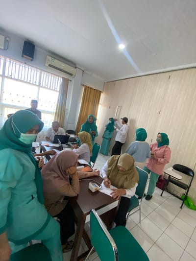 In a room, a group of people are gathered around tables. One person in the foreground is wearing a teal hijab and a teal uniform, with a surgical mask. To their right, another person in a mauve hijab is seated at a table, looking down at something. Next to them, another person in a mauve hijab is seated and writing on a paper.
In the background, a man wearing a white shirt and a black cap is standing near a window, holding a white paper. To his right, two women are standing. One is wearing a teal hijab and a teal outfit, while the other is wearing a pink jacket over a teal outfit. They appear to be interacting with each other or with a whiteboard on the wall.
Further back, a man in a white shirt is seated at a table with a laptop. Another woman in a teal hijab is seated at a table, looking at her phone. The room has white walls and a tiled floor. There is an air conditioning unit on the wall and a window with blinds. There are several chairs and tables visible. The scene suggests a training session or a meeting. The presence of medical attire on some individuals suggests a healthcare-related context. The overall atmosphere is one of focused activity and interaction.