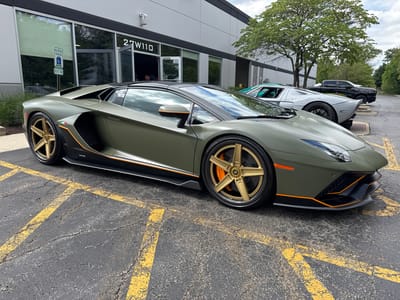 The image shows a matte olive green Lamborghini Aventador SVJ parked in a cracked asphalt parking lot marked with yellow lines. The car has gold-colored wheels and orange accents on the side skirts and front splitter. Behind the Lamborghini is a silver Ford GT and a black pickup truck. The building in the background has the address "27W110" displayed above the windows. A "Reserved Parking" sign is visible near the building entrance. A tree with green leaves is present to the right of the building. A person is partially visible in the building doorway. The scene is located in West Chicago, United States.