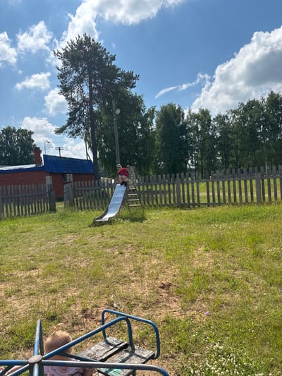 A child is sliding down a metal slide in a grassy yard. The child is wearing a red shirt and shorts, and a hat. The slide is on a playground structure with wooden steps. In the background, there is a wooden fence, a brick building with a blue roof, and trees. The sky is blue with white clouds. There is also a merry-go-round in the foreground, with a baby on it. The scene appears to be in a rural area, possibly a backyard or a small park. Based on the provided context, this scene is likely located in the city of Kurgan, Russia.