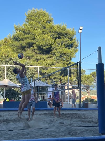 In Gošići, Montenegro, on a sunny day, a beach volleyball game is in progress on a sand court. A male player in a light-colored tank top and yellow shorts is in mid-air, reaching to hit a volleyball. He is the primary subject, captured in the act of serving or spiking. Other players are visible on the court, some in athletic attire, observing the play. The court is bordered by a net, and beyond it, trees and buildings are seen in the background. There are also beach umbrellas and floodlights, suggesting the location is set up for recreational activities. The overall scene depicts a casual, outdoor sporting event.