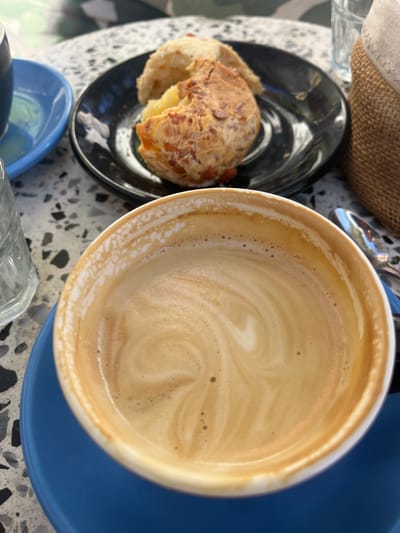 A high-angle shot reveals a cup of coffee on a blue saucer, alongside a black plate with a partially eaten pastry, all resting on a terrazzo table in Olivos, Argentina. The coffee cup is white with a tan interior, showing swirling latte art and small bubbles. The pastry on the black plate appears to have cheese baked on top and a white filling. The terrazzo table has a mix of black, white, and gray fragments. A woven basket and a glass are partially visible in the background.