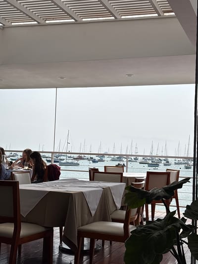 The image depicts an outdoor dining area of a restaurant with a view of a harbor filled with numerous sailboats. In the foreground, several tables are set with white tablecloths and chairs. Some people are seated at one of the tables, engaged in conversation and eating. The setting appears to be the restaurant "La Punta" in La Punta, Peru, given the presence of a marina and sailboats, a common sight in this coastal district known for its nautical activities. The overall atmosphere suggests a leisurely meal with a scenic backdrop.