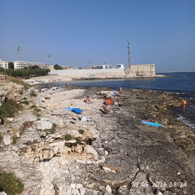 The image captures a rocky coastline near Molfetta, Italy, with several individuals enjoying the sunny weather. In the foreground, the rough, light-colored rocks are dotted with sparse vegetation. Several people are visible on the rocks, some sunbathing on towels and loungers, while others are near the water's edge.
Further back, a small sandy beach is visible, with more people swimming and relaxing. In the background, a long, modern-looking building or structure runs along the coastline, with a tower equipped with antennas. Trees and residential buildings are situated behind this structure, suggesting the proximity of an urban area. The sea is calm and blue, reflecting the clear, cloudless sky. The timestamp "23.06.2026 08:34" is superimposed on the lower right corner of the image.