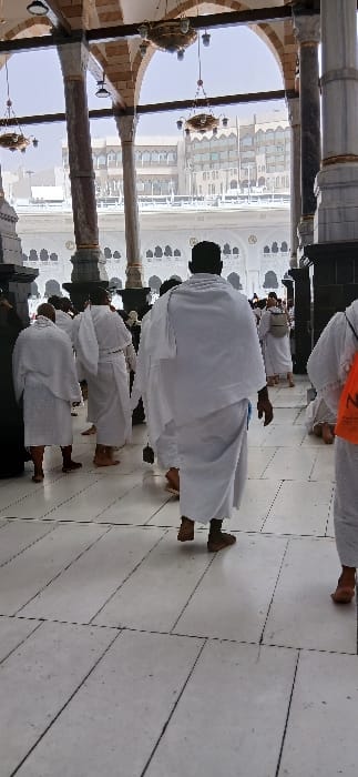 The image depicts an arcaded walkway within the Grand Mosque (Masjid al-Haram) complex in Makkah Al Mukarramah, Saudi Arabia. The ground is composed of light-colored, possibly marble or tiled, flooring with visible dark grout lines. Overhead, a series of architectural arches are supported by fluted columns of varying dark and light finishes. Multiple ornate brass chandeliers with numerous light bulbs hang from the arched ceilings.
Numerous individuals, predominantly adult males, are present, most of whom are dressed in white Ihram attire, indicating their status as pilgrims. The majority are barefoot, consistent with Ihram regulations. In the central foreground, a male figure is observed from behind, walking forward, barefoot, with a small dark object suspended from his right hand. To his left and right, several other males in Ihram are also walking. On the far right, a pilgrim wears an orange bag or cloth over their shoulder. In the mid-background, another pilgrim is visible with a light-colored backpack.
Through the arches, the exterior environment reveals modern multi-story buildings with numerous windows, typical of the urban landscape surrounding the Grand Mosque. A white, decorative balustrade or railing is visible between the arcaded area and the distant buildings, all under a bright sky. The scene shows movement and gathering within a structured religious environment.