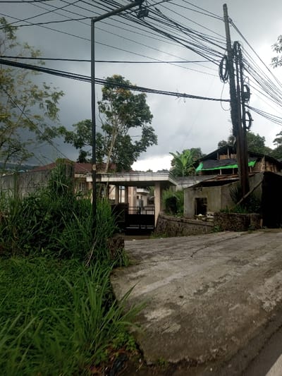 The image displays an outdoor scene in Kandapola, Sri Lanka, under a heavily overcast sky dominated by dark grey clouds. The foreground consists of a concrete road surface, exhibiting visible cracks and appearing damp, with abundant tall green grass and weeds growing along its left edge and lower left corner.
In the midground, a dark, closed metal double-leaf gate is positioned within a light-colored concrete entrance structure featuring a flat roof. Beyond this gateway, a multi-story building with light-colored walls and a brown roof is partially visible. To the left of the gate, another building with a reddish-brown roof is partially obscured by trees.
Two utility poles are present in the upper half of the image. A slender, dark utility pole stands centrally on the left, supporting multiple horizontal cables that traverse the upper frame. On the right, a sturdier utility pole is heavily laden with numerous coiled black cables and additional horizontal lines extending across the upper right quadrant.
Adjacent to the road on the right, a small, open concrete block structure, possibly derelict, with a rectangular opening, is situated behind a low stone retaining wall. Further back on the right, an unfinished or partially constructed building with exposed wooden framing and green tarpaulin coverings is visible on elevated terrain. Dense green foliage, including several large trees, is prevalent throughout the background and midground. No individuals are depicted in the scene.