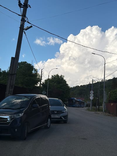 The image shows a street scene with several vehicles and infrastructure. In the foreground, two cars are parked on the right side of the road. The car on the left is dark colored and appears to be a compact hatchback. The car on the right is a lighter color and its license plate reads "H100YC 193". Further down the road, a blue bus is visible. The street is lined with trees on both sides, and a utility pole with wires and a light fixture is prominent on the left. In the background, there are hills covered in dense green vegetation. The sky is blue with large, fluffy white clouds. Two traffic signs are visible on the right side of the road: one indicates a speed limit of "35T" and the other reads "300m". Another sign with a red circle and a diagonal line through a white circle is also present. The overall setting appears to be a residential or semi-rural area.