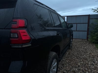 The image shows the rear passenger side of a black SUV parked on a gravel driveway. The car's taillights, tinted windows, and running boards are visible. The vehicle is covered in raindrops, suggesting it is raining. Behind the car, a grey corrugated metal fence with red metal framing stretches across the scene. To the right of the fence, a dark green evergreen tree is partially visible. The sky is overcast and appears grey. The image is set in Shebarshino, Russia.