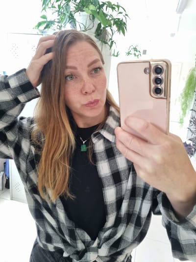 A fair-skinned woman with reddish-blonde hair and freckles is taking a mirror selfie in what appears to be an indoor setting. She is wearing a black t-shirt with a black and white plaid shirt unbuttoned and tied at the waist. She has a green pendant necklace. Her right hand is raised, touching her hair, and her left hand is holding a rose-gold colored smartphone to take the photo. Her lips are pursed. The background shows a white wall, some cabinets, and lush green plants on a shelf. There is no indication of the city of Kemul, Russia in the image.