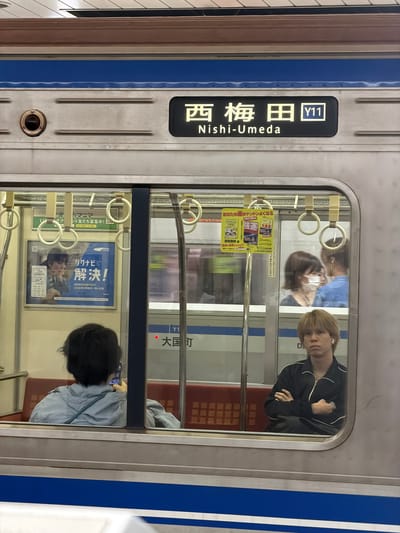 The image captures the side of a modern train car in Osaka, Japan, likely at a station platform, with its destination sign prominently displayed and the interior visible through a large window. The train's exterior is characterized by a silver-grey metallic finish accented with a distinct blue horizontal stripe. A rectangular black display panel on the train's exterior shows the destination in white text as "西梅田" (Nishi-Umeda) along with the line identifier "Y11".

Through the reflective train window, the interior elements and several passengers are visible. A young person with blond hair, wearing a dark track jacket with white trim and earbuds, is looking directly towards the camera, seemingly out of the window. Further inside, another person with dark hair, seen from behind, is focused on a mobile phone while wearing a light blue jacket. Other blurry passengers, including one wearing a face mask, are also visible in the background.

Inside the train, white circular grab handles are suspended from above, and various advertisements adorn the interior walls. One notable poster advertises "リクナビ" (Rikunabi), a job search service, with the prominent text "解決!" (Solution!) and details about "WEBの合同説明会で注目企業の情報を一気に収集" (Gather information on interesting companies at the online joint company briefing). Another advertisement promotes "あなたの運がドンドンよくなる" (Your luck will steadily improve) and "強運" (Good Fortune), mentioning "深見東州" (Toshu Fukami) and "194万部" (1.94 million copies). Etiquette signs reminding passengers to "つっておすわりください" (Please hold on and sit down) and to ensure "できるだけ多くの方にご利用いただけるよう" (as many people as possible can use it) are also discernible. Reflections in the window also show faint text for "大国町" (Daikokucho) and "Y16", likely another station on the line. The scene appears to take place during the daytime.