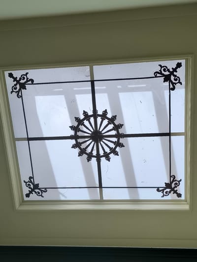 The image shows a ceiling skylight, likely an interior shot. The skylight is rectangular with a white frame. Dark metal decorative elements are overlaid on the glass: corner flourishes and a central, wheel-like design. The glass appears to be translucent, allowing diffused light through. The room beyond the skylight is not clearly visible due to the light diffusion. The location is not discernible from the image itself. It is impossible to determine if this is in Utrecht, Netherlands, based on the visual information provided.
