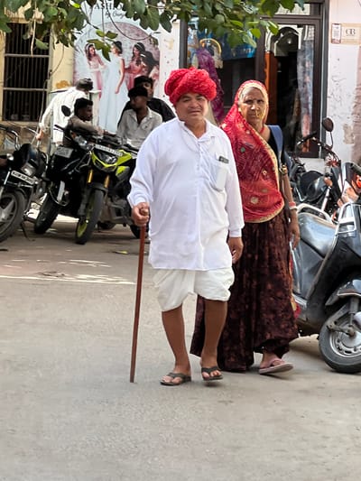 In Pushkar, India, a man and a woman stand in front of a building with a shop. The man, dressed in a white kurta and dhoti with a red turban, leans on a cane. The woman wears a red and gold sari. In the background, motorcycles are parked and a poster advertising fashion is displayed. The scene appears to be a street in a bustling Indian city.
