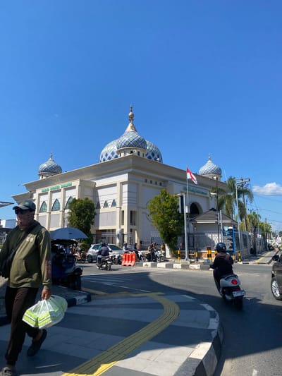 In the city of Jawa, Indonesia, under a clear blue sky, a mosque with intricate patterned domes stands prominently. The mosque, named "MASJID SYAHRIYAH SHALIM," features a white facade with decorative arches and windows. A red and white Indonesian flag flies from a flagpole outside the mosque. In the foreground, a person wearing a green hoodie, sunglasses, and a face covering walks with a plastic bag containing groceries. They are on a pedestrian crossing with yellow tactile paving. Several motorbikes and cars are present on the street, with some people riding motorbikes. Traffic cones and barriers are visible, suggesting ongoing construction or road work. Palm trees and other greenery surround the mosque, adding to the urban landscape. The scene depicts a typical day in an Indonesian city, with daily life and religious structures coexisting.
