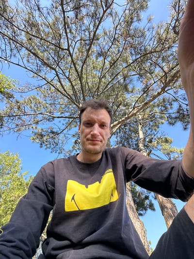 In this ground-level, wide-angle selfie, a man with short brown hair and a slight stubble is pictured from the chest up. He is wearing a black long-sleeved shirt with a large, bright yellow graphic design on the front, which appears to be a stylized smiley face with a jagged outline. The man's expression is neutral, and he is looking directly at the camera. His right hand is partially visible in the upper right corner of the frame, suggesting he is holding the camera.
The man is positioned outdoors, beneath a large pine tree whose branches and needles fill a significant portion of the upper frame. The sky behind him is a clear, vibrant blue. To the left, glimpses of other green foliage and tree trunks are visible. The setting appears to be a natural outdoor environment, possibly a park or a forested area, and it is consistent with the general landscape of Velykozalissia, Ukraine, a town known for its green spaces and surrounding natural beauty. The lighting indicates it is daytime, likely a sunny day, given the bright blue sky and the illumination on the man and the trees.