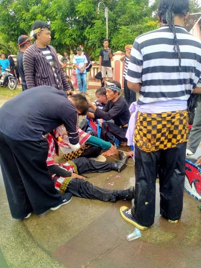 The image shows a scene in Kecamatan Kedawung, Indonesia (latitude: -7.52, longitude: 111.029).  There are several men standing and sitting on a concrete surface, surrounded by green foliage. A man in a black shirt is crouched down, apparently tending to a man wearing a white and red shirt who is sitting on the ground with his legs stretched out.  There are multiple men and women in the background. One man is wearing a black and white striped shirt and black pants, and a checkered sash around his waist. There are other men around him, and a few women are visible in the background.  The man in the checkered sash is looking down at the men in the foreground.  There is a discarded plastic cup on the ground, and a few pink flowers are scattered on the concrete surface.  The image is likely a snapshot of a traditional ceremony or event taking place in the town.