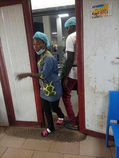 Two individuals, likely kitchen staff, walk through a doorway. The first person, a woman, wears a light blue hairnet and matching top with dark pants and open sandals. Her face mask is pulled down. Behind her, a taller person with a similar hairnet wears a white shirt, dark green apron, and maroon pants with dark sandals. The doorway appears to lead into a larger room, possibly a cafeteria, with visible tables and chairs. A "Gold Custard" package sticker is visible on the door frame. The floor has dirty water stains.