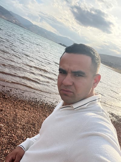 A medium shot, canted to the right, shows a man in a white, long-sleeved shirt standing on a pebble beach next to a large body of water. Mountains are visible in the background under a cloudy sky. The man faces the camera with a neutral expression. The scene is set in Bin El Ouidane, Morocco, as implied by the landscape typical of the region.