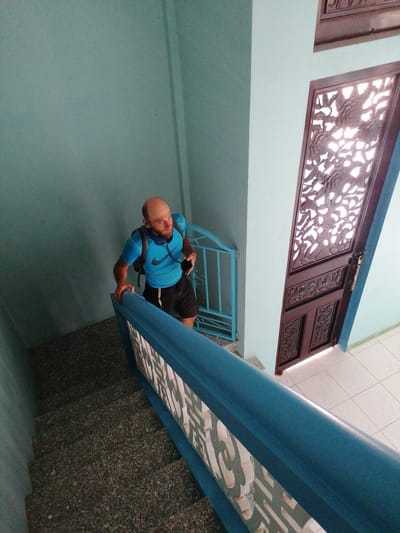 A bald, bearded man wearing a blue t-shirt, black shorts, and a black backpack walks up a granite staircase. He is holding a black smartphone in his right hand and has earbuds in his ears. The staircase has a blue railing with a decorative white insert. The walls are painted a light blue. To the right of the staircase, there is a brown wooden door with intricate carvings and a light blue metal gate. The scene appears to be an interior staircase in Lộc Quý, Vietnam.