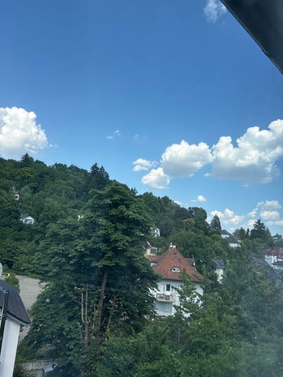 The image captures a view from a high vantage point, looking down on a densely wooded hillside with several houses nestled amongst the trees. The sky is a vibrant blue, dotted with fluffy white clouds, suggesting a pleasant, sunny day. The time of day appears to be late morning or early afternoon, given the bright lighting and the angle of the shadows.

The hillside is covered in lush green foliage, with various types of trees creating a dense canopy. Several buildings are visible, characterized by their pitched red-tiled roofs and white or light-colored walls. Some have balconies or multiple windows. The architectural style suggests a European setting, consistent with the provided location context of Schwäbisch Gmünd, Germany.

In the foreground, a dark, solid object, possibly a railing or a structural element, partially obscures the view, indicating that the photo was taken from a balcony, window, or elevated platform. Below the hillside, a paved area, likely a road or parking lot, can be seen with a light-colored car parked on it. Power lines are also visible, strung between trees, a common sight in residential areas.

The overall scene evokes a sense of tranquility and a connection with nature, despite the presence of human habitation. The clear sky and abundant greenery contribute to a peaceful and serene atmosphere. There is no visible text in the image.