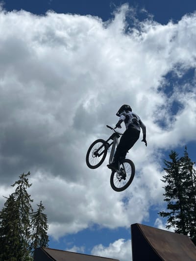 A silhouetted mountain biker in a helmet and a white and black patterned jersey and black pants is captured mid-air, performing a jump against a cloudy blue sky. The biker is on a bicycle, suspended in the air, with the front wheel higher than the rear. The background features tall evergreen trees and a blue sky with white and gray clouds. In the foreground, a portion of a wooden ramp is visible, suggesting a ski resort or a designated biking area. The image appears to be taken in a mountainous or forested outdoor location, likely in the city of Samokov, Bulgaria, known for its ski resorts and natural beauty.