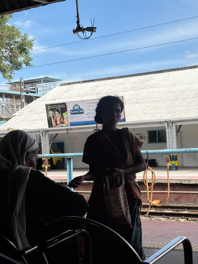 This is a photograph taken in Thanjavur, India, featuring two individuals. The primary subject is a person with dark hair and skin, wearing a brown garment, seated on a chair and looking towards the left. They are holding a patterned cloth in their hands. Behind this person, a yellow object and a blue pipe are visible. A sign board with text in Tamil is also present. The background includes a building with a corrugated roof and a clear blue sky. There are electrical wires running across the sky.
In the foreground, another person is partially visible, wearing a light-colored head covering and dark clothing. They are seated and appear to be resting or looking down. The overall setting suggests an outdoor or semi-outdoor location, possibly a street or a waiting area, with the bright sky indicating daytime. The light is strong, suggesting a sunny day.
The visible Tamil text on the sign board, partially transcribed as "சிறிய சேமிப்பை மேலும் காப்பீட்டையும் பெறுங்கள்", translates to "Get small savings and more insurance." This suggests the sign is an advertisement for financial services. The image captures a moment of everyday life in Thanjavur, highlighting the presence of people and local commerce.