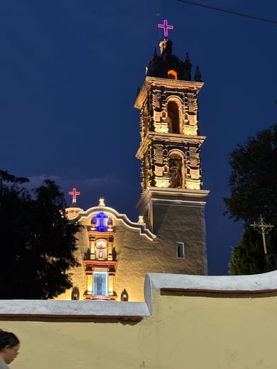 The image depicts a church in Cholula, Mexico at night. The church has a tall, ornate bell tower lit up in warm yellow light, topped with a bright pink cross. The sky is dark blue. The church facade is painted pale yellow and is illuminated with similar lighting. The building has a central window framed in red and blue light above the main entrance, topped with a blue cross, with trees in the foreground and to the side, obscuring the lower portions of the church. The image is turned sideways. A secondary building is seen with a decorative metal cross on the top right.