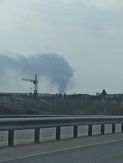 The image shows a view from a bridge or elevated structure in Kitsjanovo, Russia. Visible in the foreground is a section of the bridge's railing. In the middle distance, a construction crane is silhouetted against a cloudy sky. Plumes of smoke are visible behind the crane, possibly indicating an industrial area or event. A landscape with a church or tall building on a small hill is visible further back.
