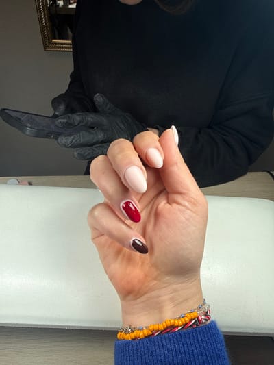 The image depicts a close-up view of a person's left hand and lower forearm within a nail salon in Aix-en-Provence, France. The hand exhibits manicured, almond-shaped nails; the thumb, index, and middle fingers are coated in a light nude polish, while the ring finger is dark red with a white line at the lunula, and the pinky finger is dark brown with a matching white lunula line. The wrist is adorned with multiple bracelets, including orange beaded strands, a thin silver chain with small blue beads, and a woven bracelet in red, white, and black. The forearm is covered by a royal blue long-sleeved garment. In the background, partially obscured, a second individual, likely a nail technician, is visible, wearing a black long-sleeved top and black protective gloves. This individual is holding a dark-colored smartphone horizontally in their gloved hands. The client's hand is positioned above a white, rectangular padded armrest, which rests on a light-colored wooden surface. A gold-framed mirror is affixed to a dark wall in the upper left portion of the background.