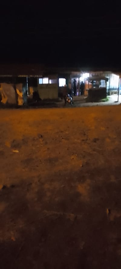 At night, a blurry scene reveals a building with glowing windows and a makeshift shelter. People gather near the entrance, while a motorcycle sits parked nearby. The foreground is a dirt area, textured with uneven ground and small debris. A dark, starless sky fills the upper portion of the image, creating a stark contrast to the lit areas below.