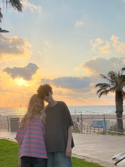 The image captures a coastal scene at either sunrise or sunset, likely in Herzliya, Israel. In the foreground, two young individuals pose affectionately. A young woman with long blonde hair, wearing a pink and grey striped t-shirt, a black choker, and visible braces, smiles at the camera while leaning into a young man. The man, with dark, spiky hair, sunglasses, a dark grey t-shirt, and blue jeans, has his arm around her shoulder, and his head gently rests against hers. Behind them, the sun, low on the horizon and partially obscured by clouds, casts a golden glow across the Mediterranean Sea and the sky. A small white sailboat is visible on the water in the distance. A sandy beach with indistinct figures is separated from a paved promenade by a chain-link fence, which features a blue and white sign with Hebrew text. A large palm tree stands on the right, and green grass borders the paved area in the immediate foreground. A street light fixture is partially visible in the top left.