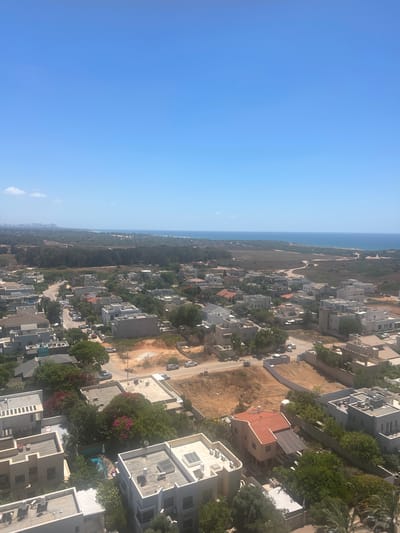 This is an aerial view of a residential area in Hadera, Israel, on a clear, sunny day. The scene is dominated by a vast expanse of blue sky, with the horizon meeting the tranquil blue sea. In the foreground, a suburban neighborhood unfolds with numerous houses, primarily featuring white and light-colored facades, flat roofs, and some with red tile roofs. The architecture appears modern, with a mix of detached homes and some multi-unit buildings. Green trees and vegetation are interspersed throughout the neighborhood, with some flowering trees displaying pink blossoms. Several cars are visible parked along the streets and driveways, indicating an active residential area. Further in the distance, beyond the immediate residential zone, a dense line of trees creates a natural buffer before the sandy landscape leading to the coastline. The sea stretches out to the horizon, and a faint city skyline can be discerned in the far distance along the coast. The overall impression is one of a peaceful, sun-drenched coastal community.